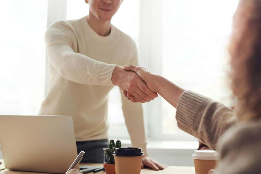 Two people shaking hands across a table with a laptop and two coffees on it.